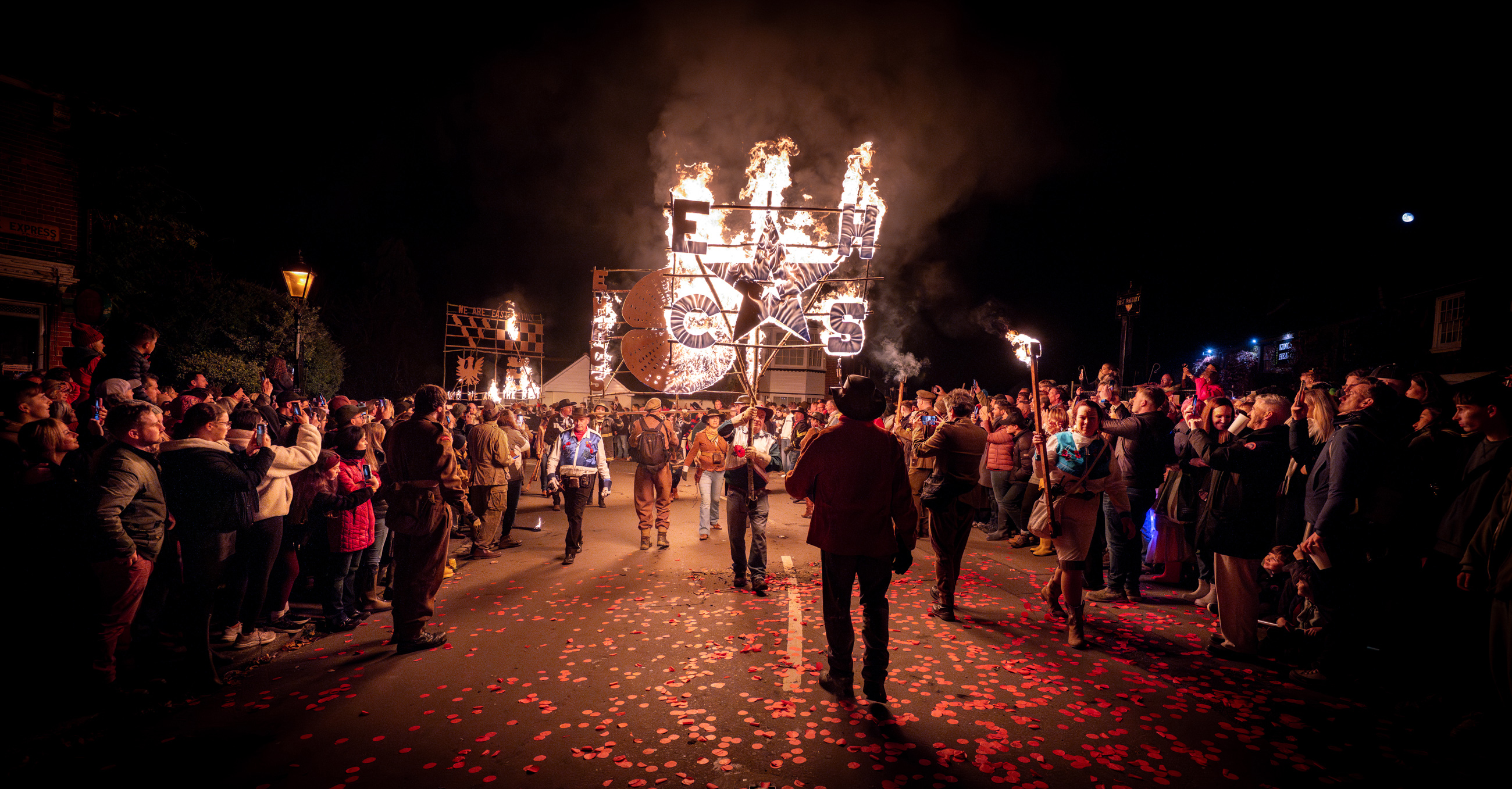 Crowds watching the East Hoathly Carnival and Bonfire parade as participants carry a large illuminated burning display through the village streets at night.